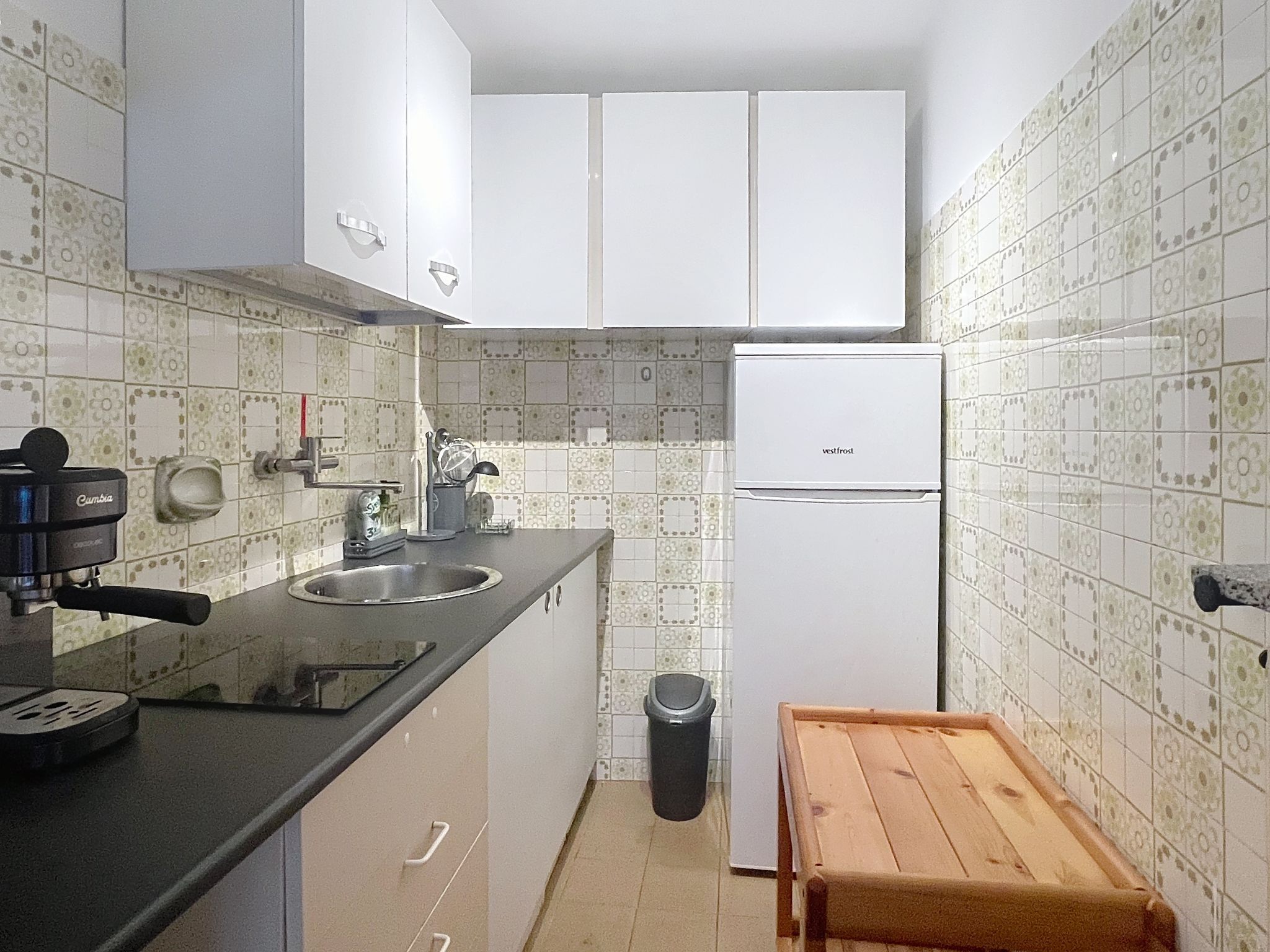 A compact kitchen with white cabinets, a black countertop, a stainless steel sink, a white refrigerator, and a wooden stool.