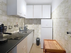 Eine kompakte Küche mit weißen Schränken, einer schwarzen Arbeitsplatte, einem Edelstahl-Spülbecken, einem weißen Kühlschrank und einem hölzernen Hocker.