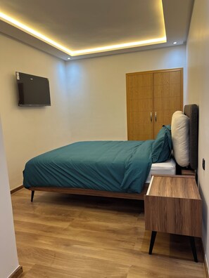 A bedroom with a bed, a wooden wardrobe, a flat-screen TV, and a bedside table. - Beautiful 2-bedroom apartment with a view of the pool (Sidi rahal)