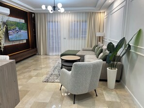 A modern living room with a sectional sofa, a round coffee table, a grey armchair, a TV mounted on a wooden panel, and a large potted plant. - Beautiful 2-bedroom apartment with a view of the pool (Sidi rahal)