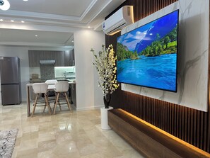 A modern living room with a flat-screen TV mounted on a wall, a dining area with a white table and chairs, and a kitchen in the background. - Beautiful 2-bedroom apartment with a view of the pool (Sidi rahal)