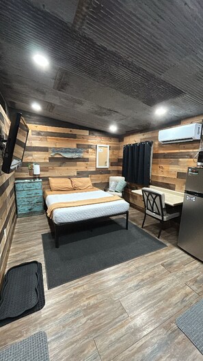 A compact room with a bed, a small desk, a chair, and a kitchenette area. - The Adventure Center (Pineville)