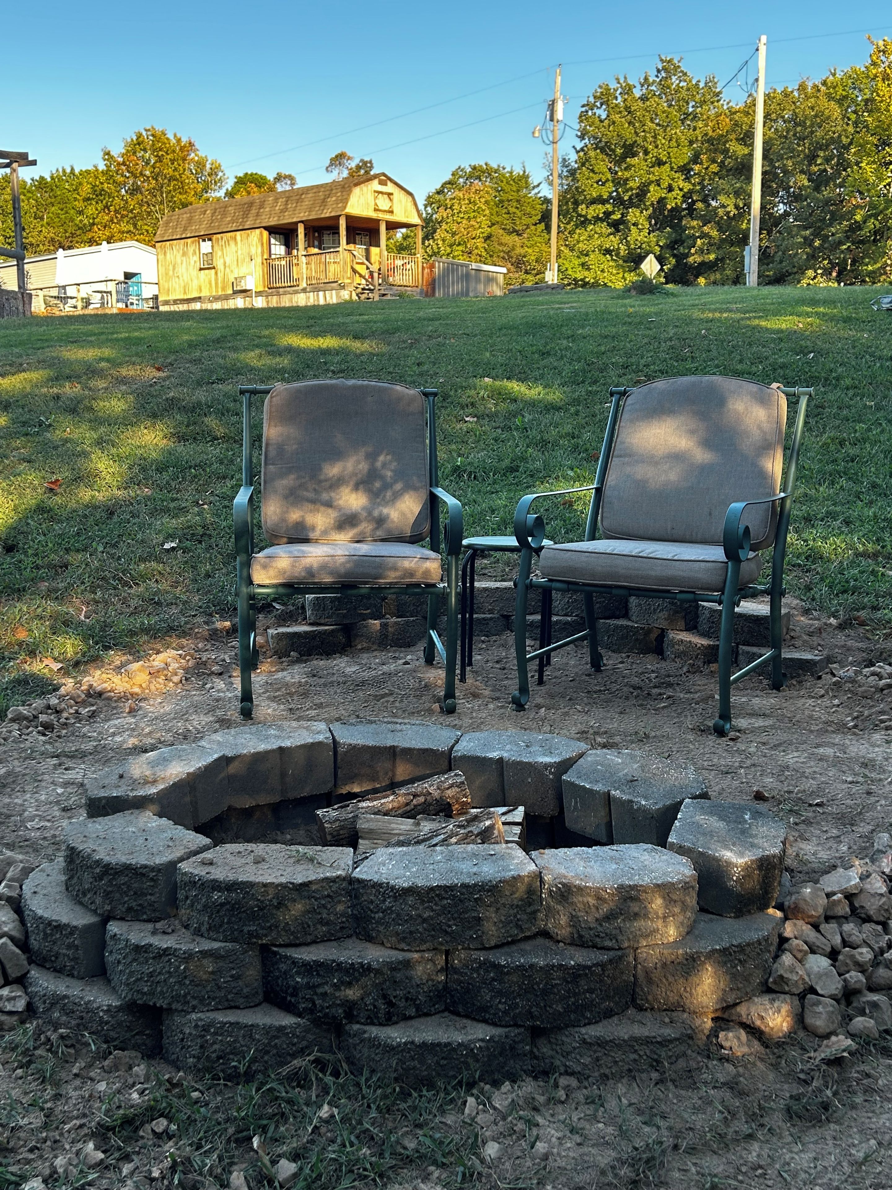 A fire pit with two chairs, surrounded by a stone fire ring, in a grassy outdoor area with a house in the background.