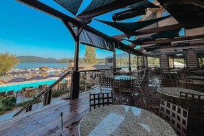 A covered outdoor seating area with tables and chairs, overlooking a swimming pool and a body of water. - Hot Tub, Fireplace, Spacious Deck & Serene Views (Whitefish)