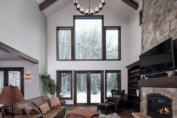 A spacious living room with a fireplace, large windows, and a stone accent wall. - Spacious Home w/ Resort Amenities l Full Kitchen, Nature Trails & Fireplace (Whitefish)