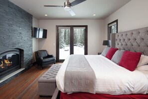 A bedroom with a fireplace, a large bed with multiple pillows, a ceiling fan, and a view of a snowy landscape through the door. - Spacious Home w/ Resort Amenities l Full Kitchen, Nature Trails & Fireplace (Whitefish)