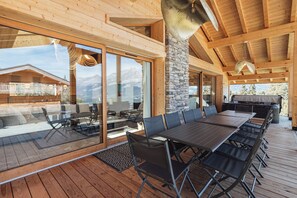 An outdoor dining area with a wooden deck, glass sliding doors, and a stone pillar. - Chalet Eteila with Sauna, Jacuzzi and Cinema (Combloux)