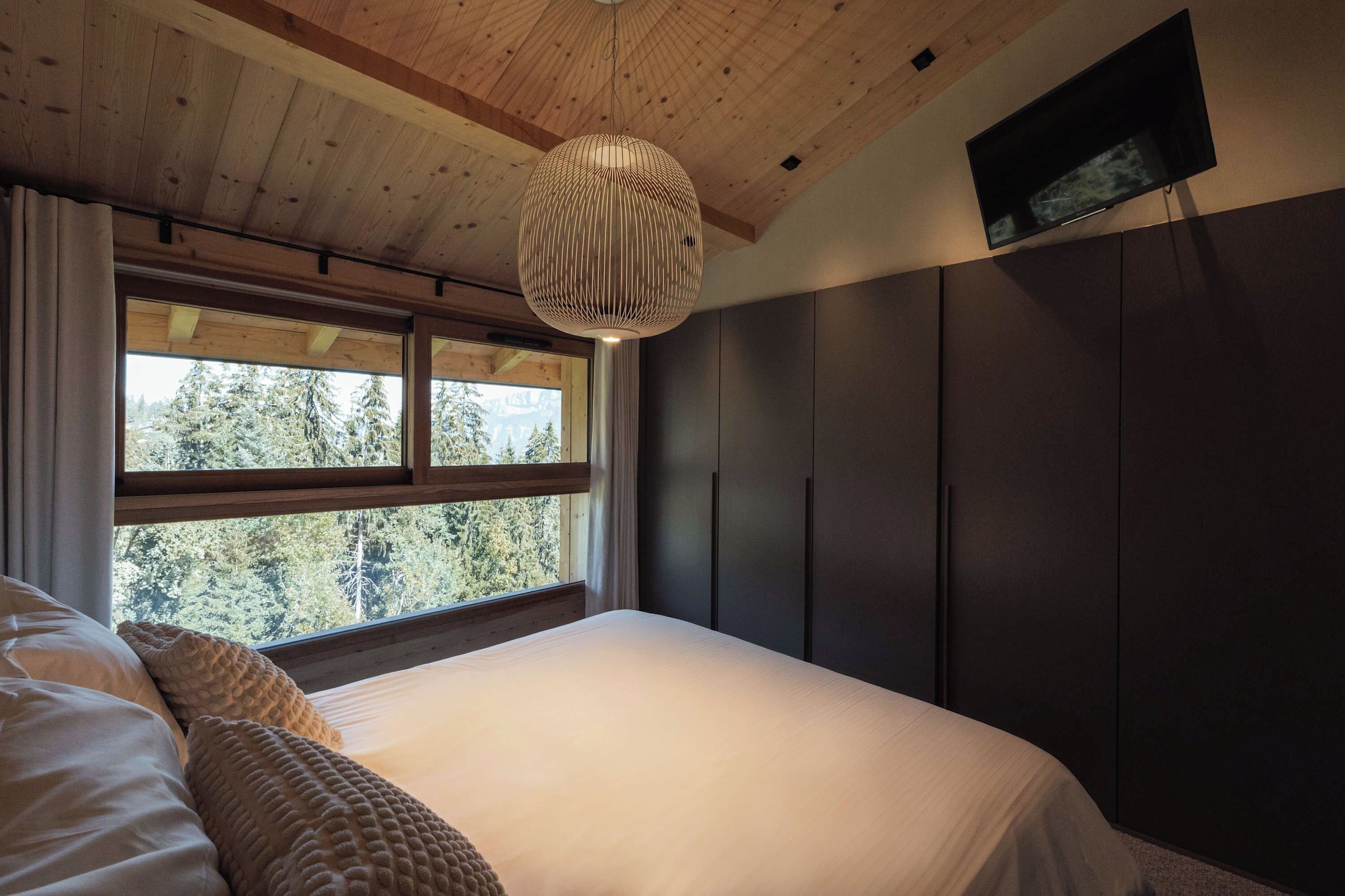 Een slaapkamer met een groot bed, een hanglamp, een aan de muur bevestigde televisie en een raam met uitzicht op bomen.