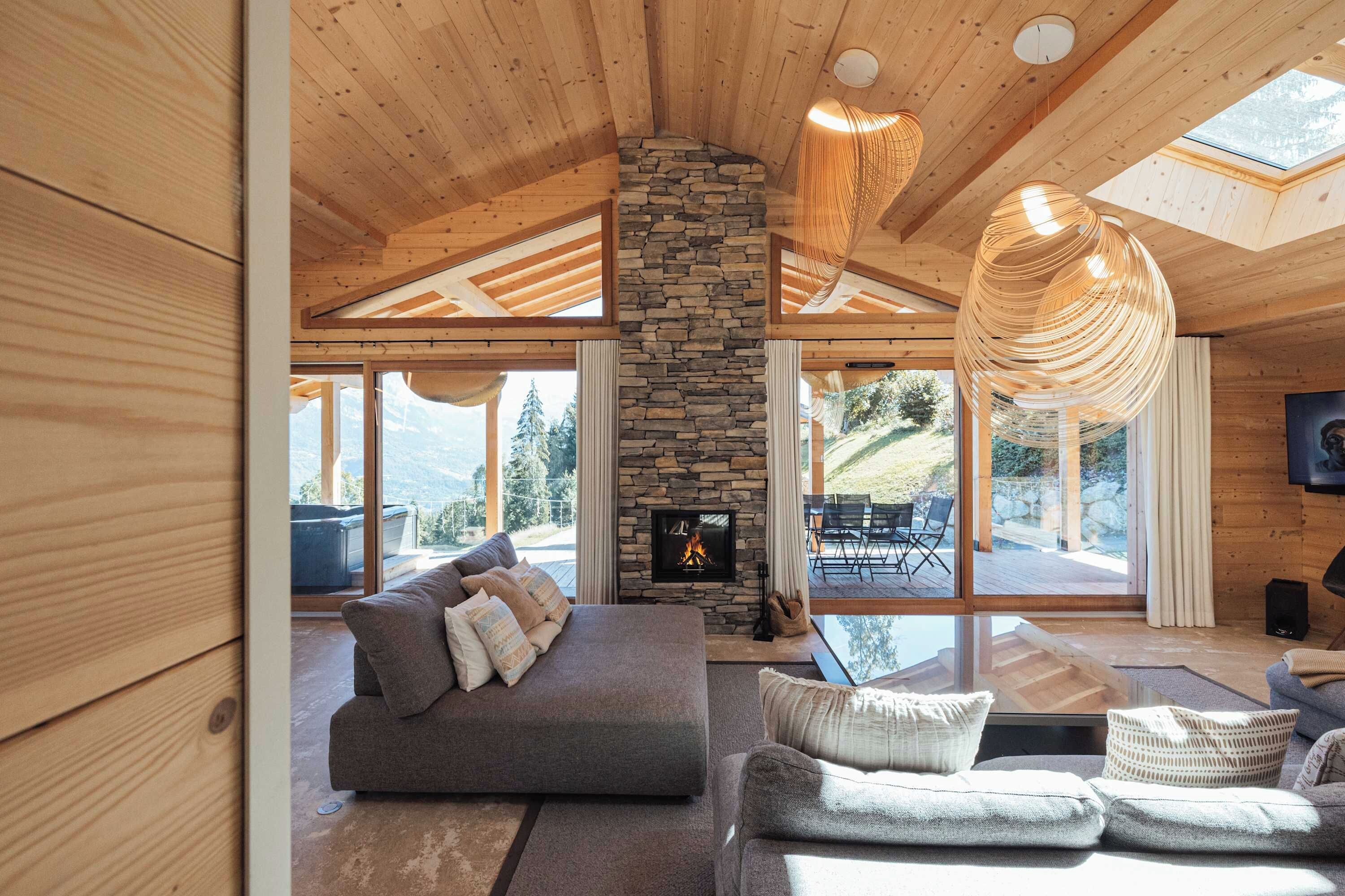 A spacious living room with a stone fireplace, wooden ceiling beams, and large windows offering a view of the outdoors.