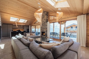 A spacious living area with a large sofa, a fireplace, and a view of the mountains through large windows. - Chalet Eteila with Sauna, Jacuzzi and Cinema (Combloux)