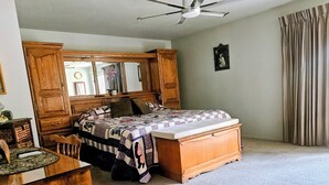 Un dormitorio con una cama, un tocador de madera, un escritorio, una silla, un ventilador de techo y una ventana con cortinas.