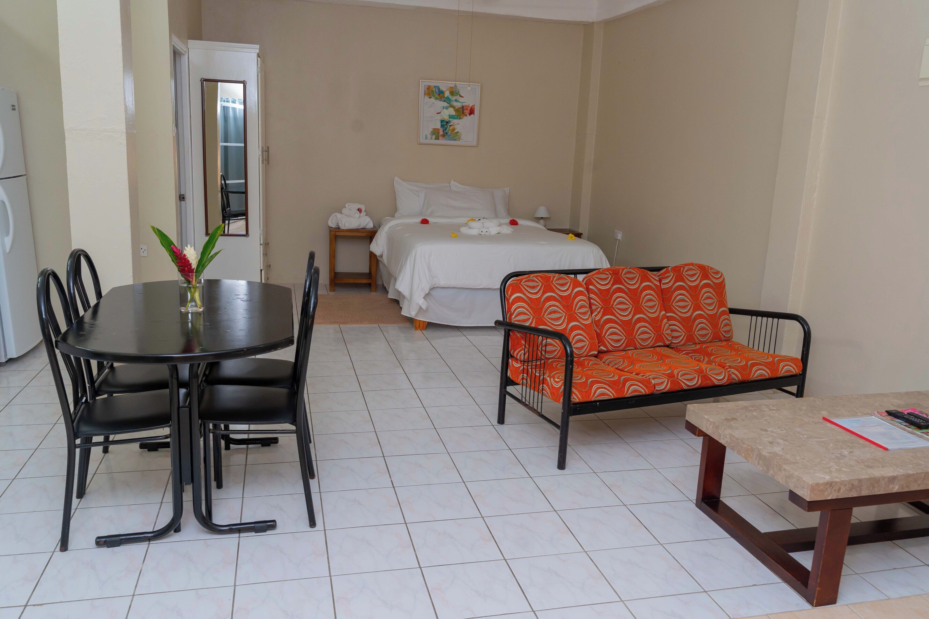 A hotel room with a bed, a sofa, a dining table with chairs, and a small table with a book and remote control.