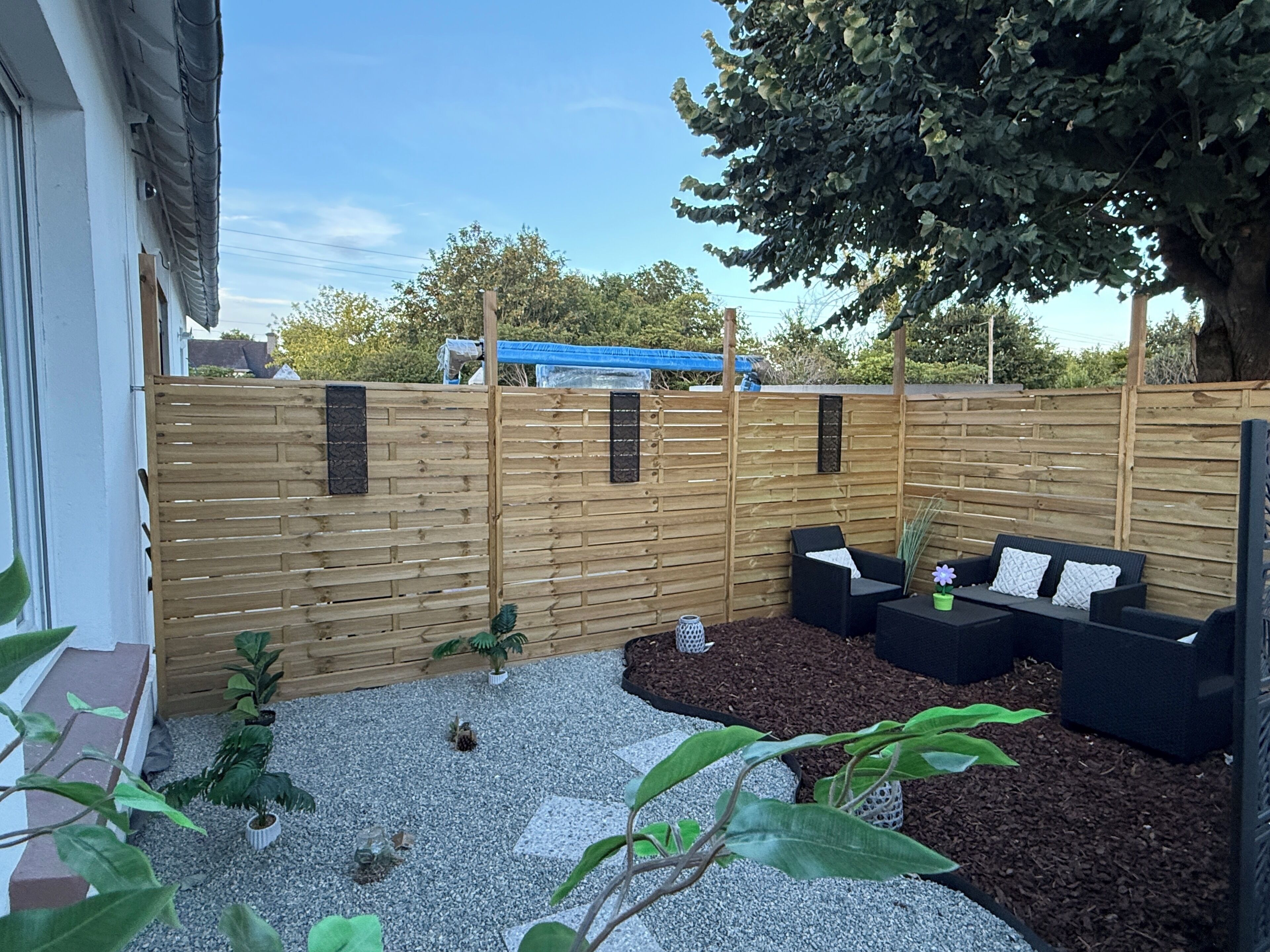Une clôture en bois, des sièges installés à l’extérieur et des plantes en pot dans un espace aménagé en jardin.