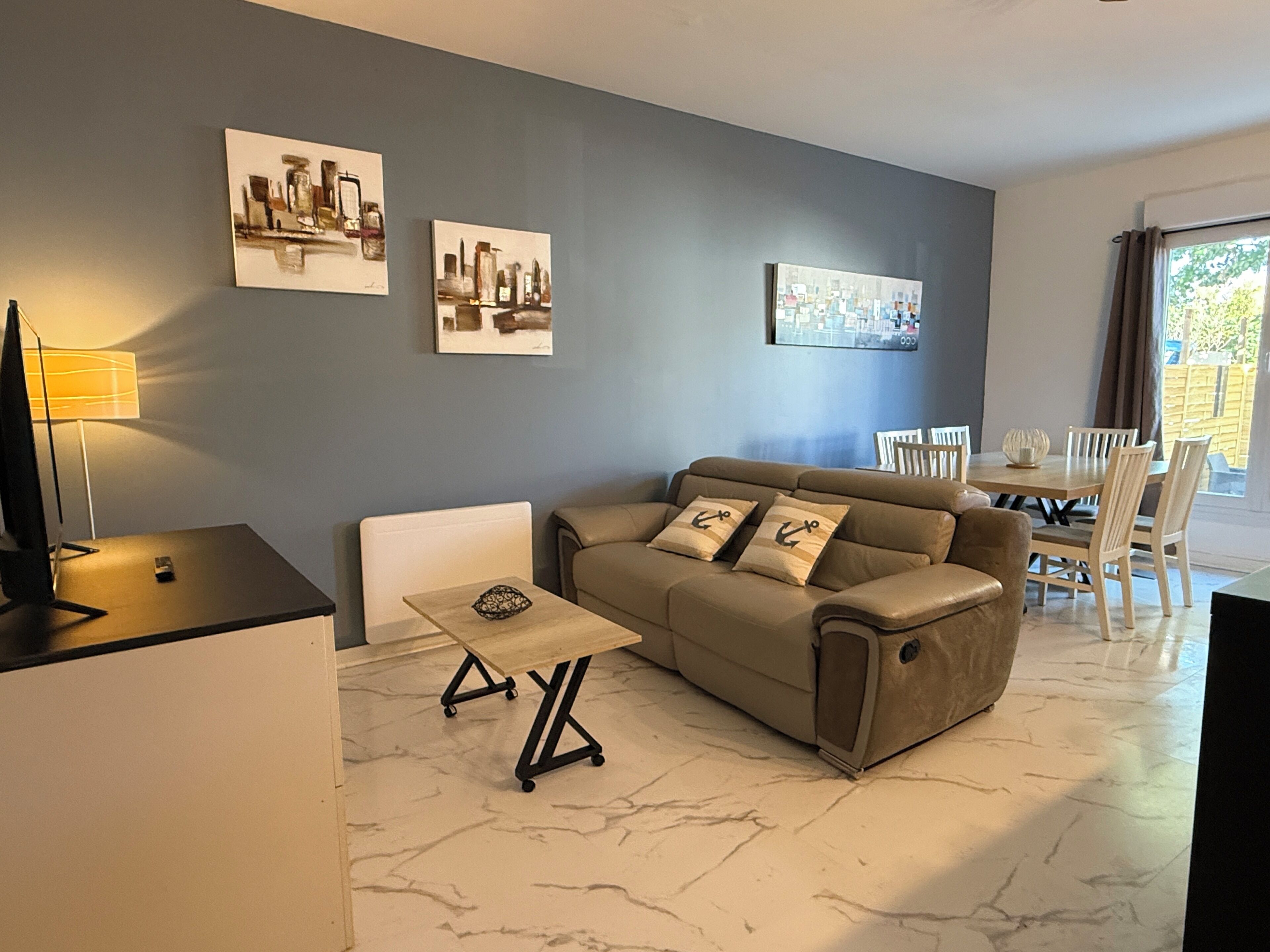 Un salon avec un canapé, une table basse et un meuble pour la télévision. Il y a trois tableaux au mur et une table à manger avec des chaises.