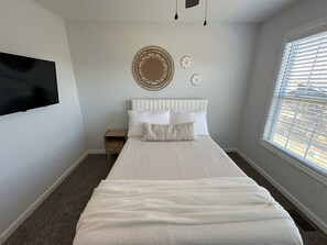 A bedroom with a large bed, a bedside table, a wall-mounted TV, and a window with blinds. - Charming 2 Bed Home near Berea College, I-75  WIFI Provided (Berea)