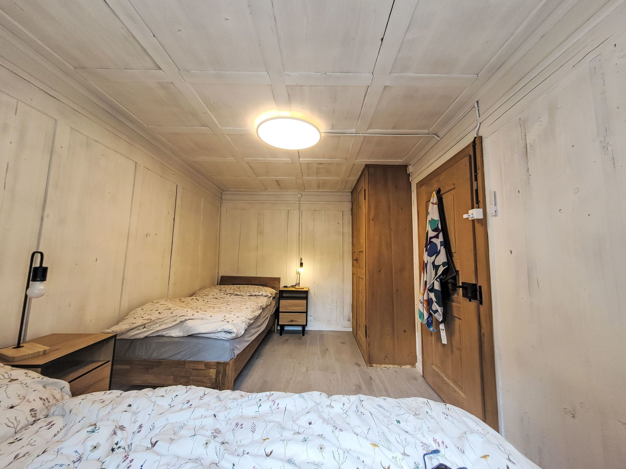 Un petit logement composé d’une seule pièce, comprenant deux lits, une table de chevet avec une lampe, une armoire en bois et un luminaire au plafond.