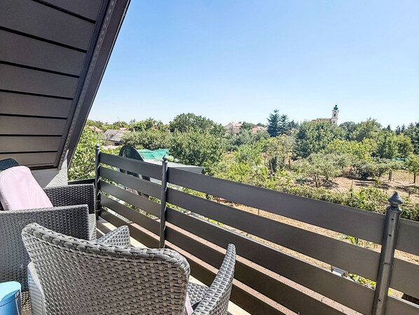 A balcony with wicker furniture and a view of a vineyard and a church tower.