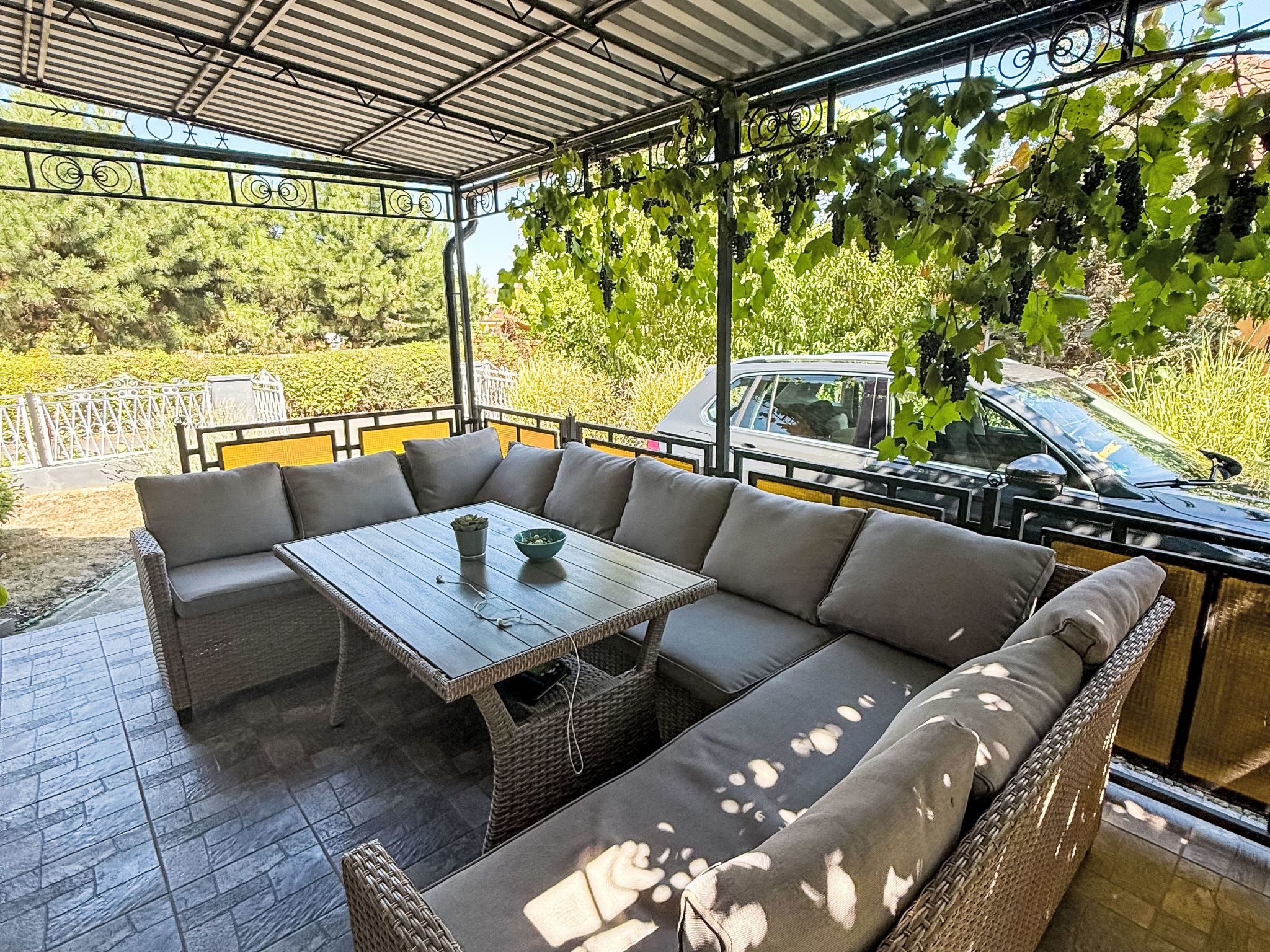 Eine Außenpatio mit Tisch und Sitzgelegenheiten, umgeben von viel Grün, mit einem Auto im Hintergrund.