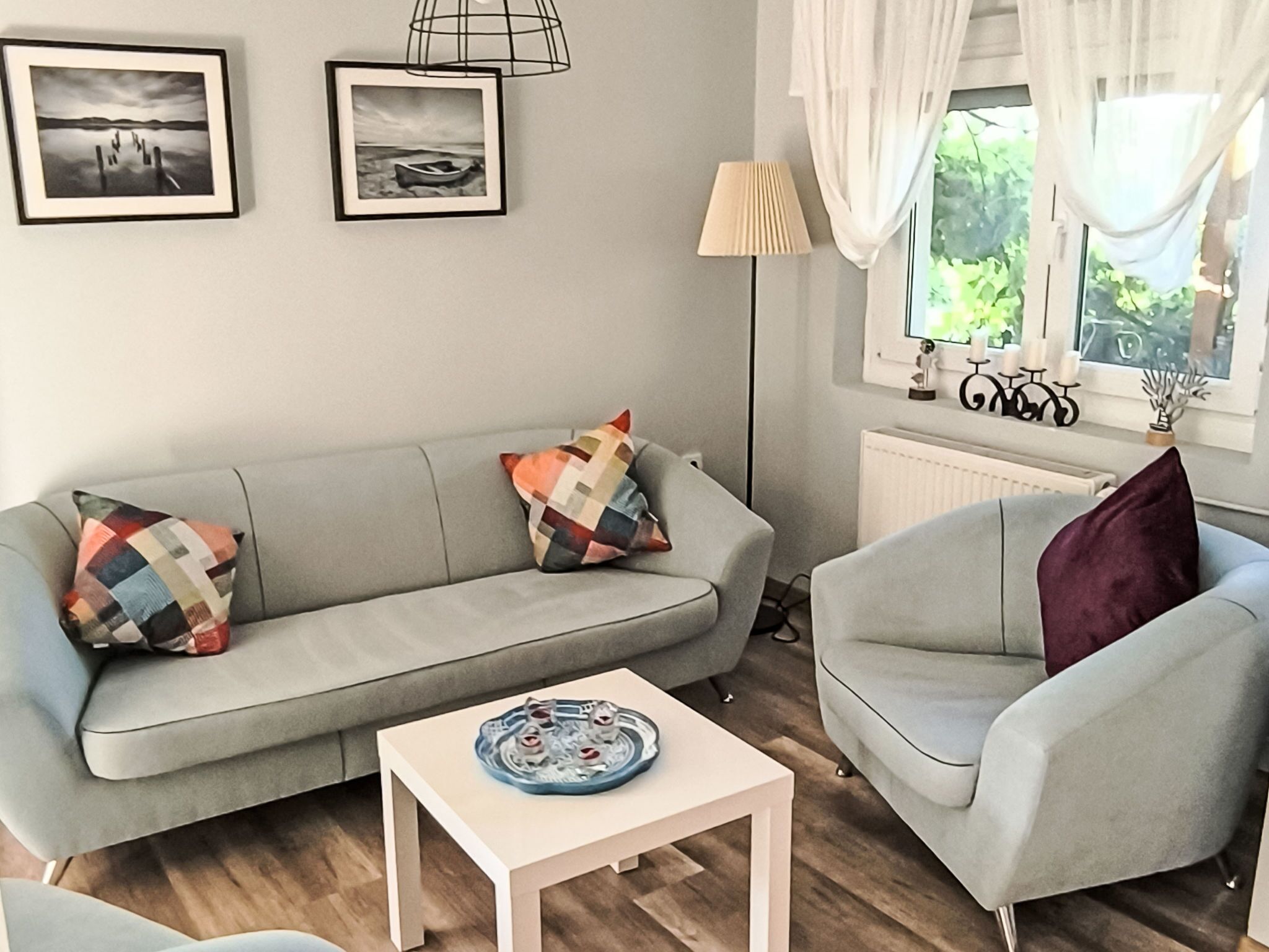 Een woonkamer met een grijze bank, een fauteuil in dezelfde kleur, een klein wit tafeltje en een raam met gordijnen.