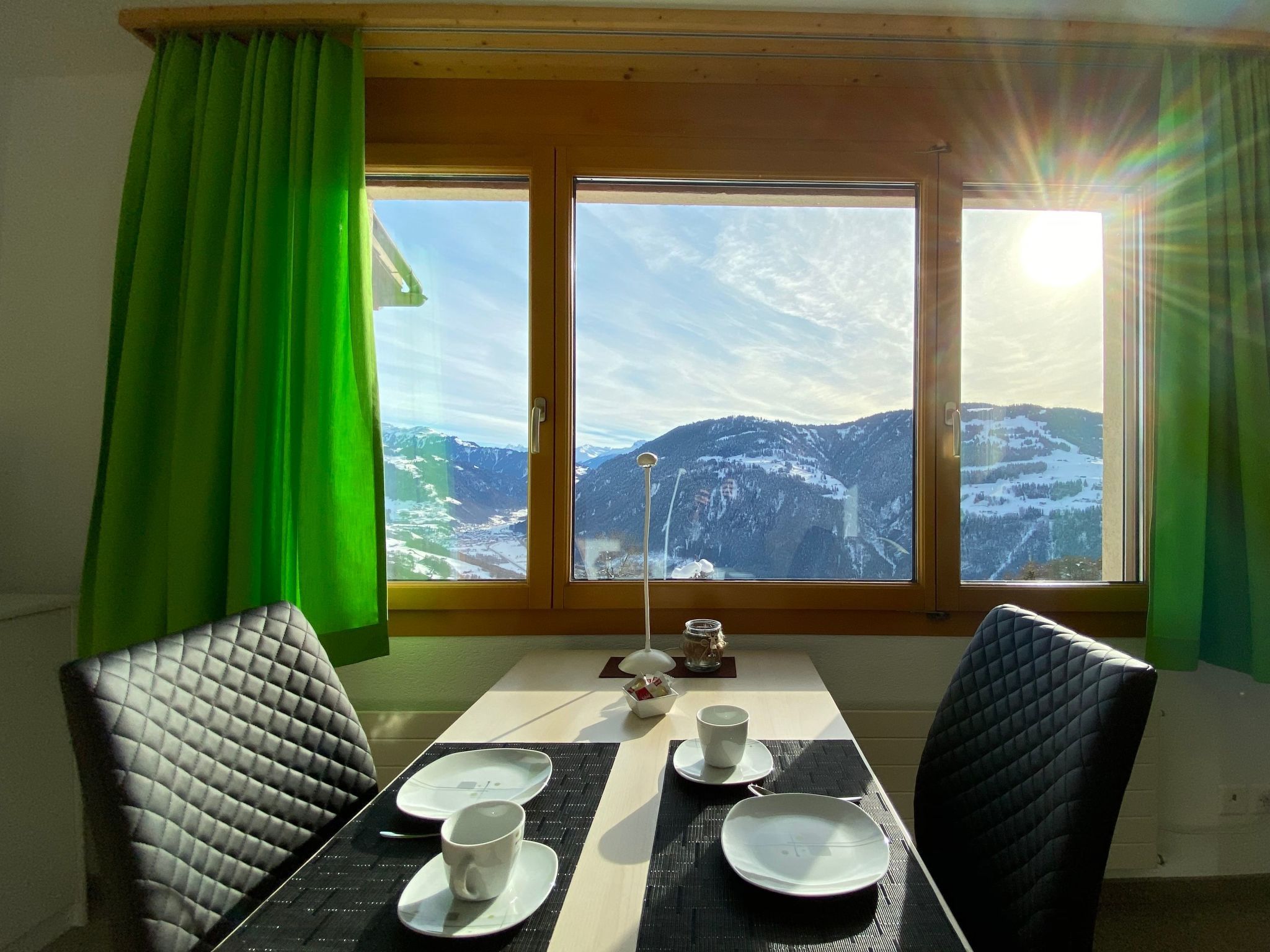 Ein Essbereich mit Tisch für zwei Personen, Blick auf schneebedeckte Berge und einem Fenster mit Vorhängen.