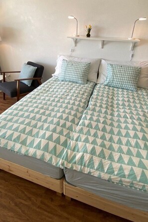 Una cama con una colcha de diseño geométrico, una mesita de noche con una lámpara y una silla en una habitación con suelo de madera.