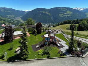 A playground with slides and climbing structures, surrounded by a grassy area and trees, with mountains in the background.