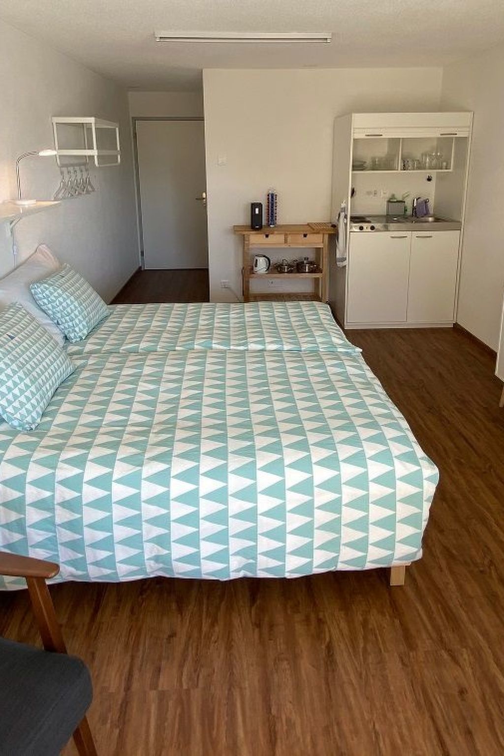 A bedroom with a bed, a wooden floor, a kitchenette, and a door.