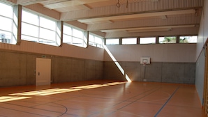 Un terrain de basket-ball intérieur doté d’un plancher en bois, d’un panier de basket et de grandes fenêtres laissant entrer la lumière naturelle.
