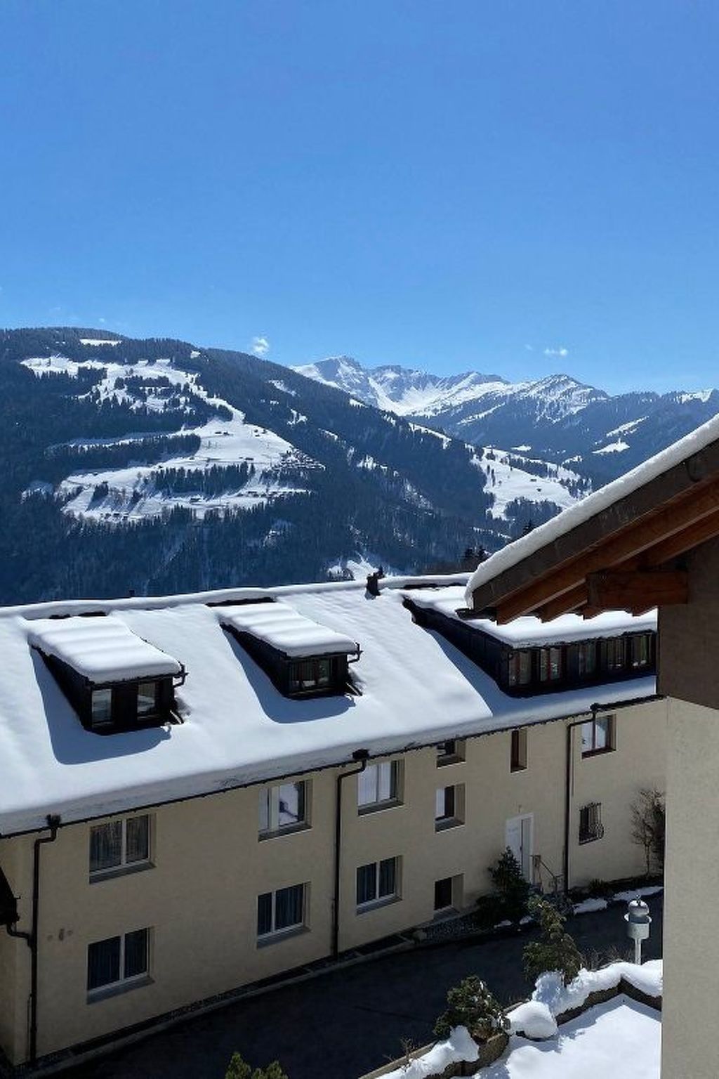 Un edificio con múltiples ventanas y un techo plano cubierto de nieve, con vistas a una cadena montañosa nevada.