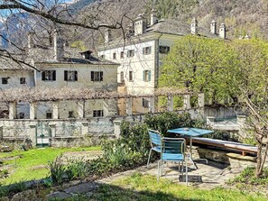 Une cour avec une table et des chaises, entourée de bâtiments aux murs de pierre, avec une chaîne de montagnes en arrière-plan.