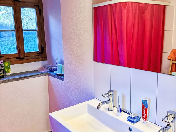 Une salle de bain avec un lavabo blanc, des robinets chromés, un rideau de douche rouge et une fenêtre donnant sur un paysage enneigé.