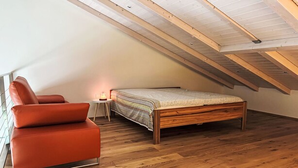 Ein Schlafzimmer mit schrägverdeckter Decke, einem hölzernen Bett, einem roten Sessel und einem kleinen Tisch mit Lampe.
