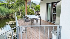 Eine Holzterrasse mit Tisch und Stühlen, die auf ein Gewässer und viel Grün blickt.
