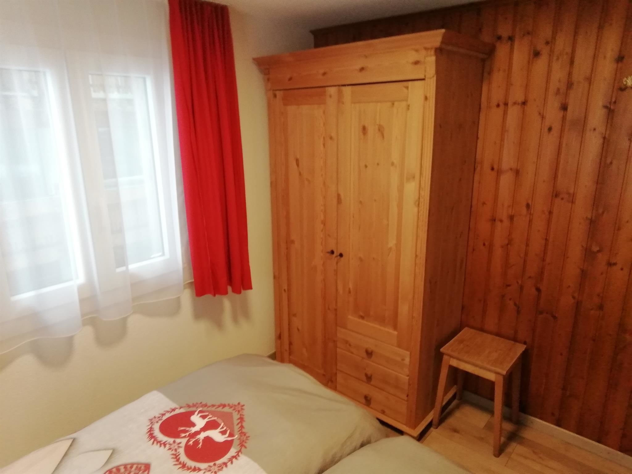 Una camera da letto con un letto, un armadio in legno, uno sgabello e una finestra con tende.