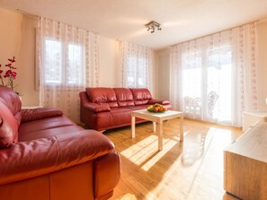 A living room with a red sofa, a wooden coffee table with fruit, and large windows with sheer curtains. - Chalet Bambi by Interhome (Saas-Fee)