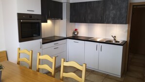 Een kleine keuken met witte kastjes, een zwart aanrecht, een inbouwoven, een spoelbak en houten stoelen.