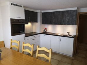 A compact kitchen with white cabinets, a black countertop, a built-in oven, a sink, and wooden chairs. - Chalet Bambi by Interhome (Saas-Fee)