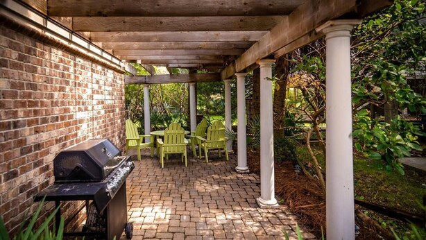 A covered patio with a brick floor, a grill, and green chairs. - 4BR Beach House w/ Covered Porches, Game Room & Shared Creek Dock Access (Pawleys Island)