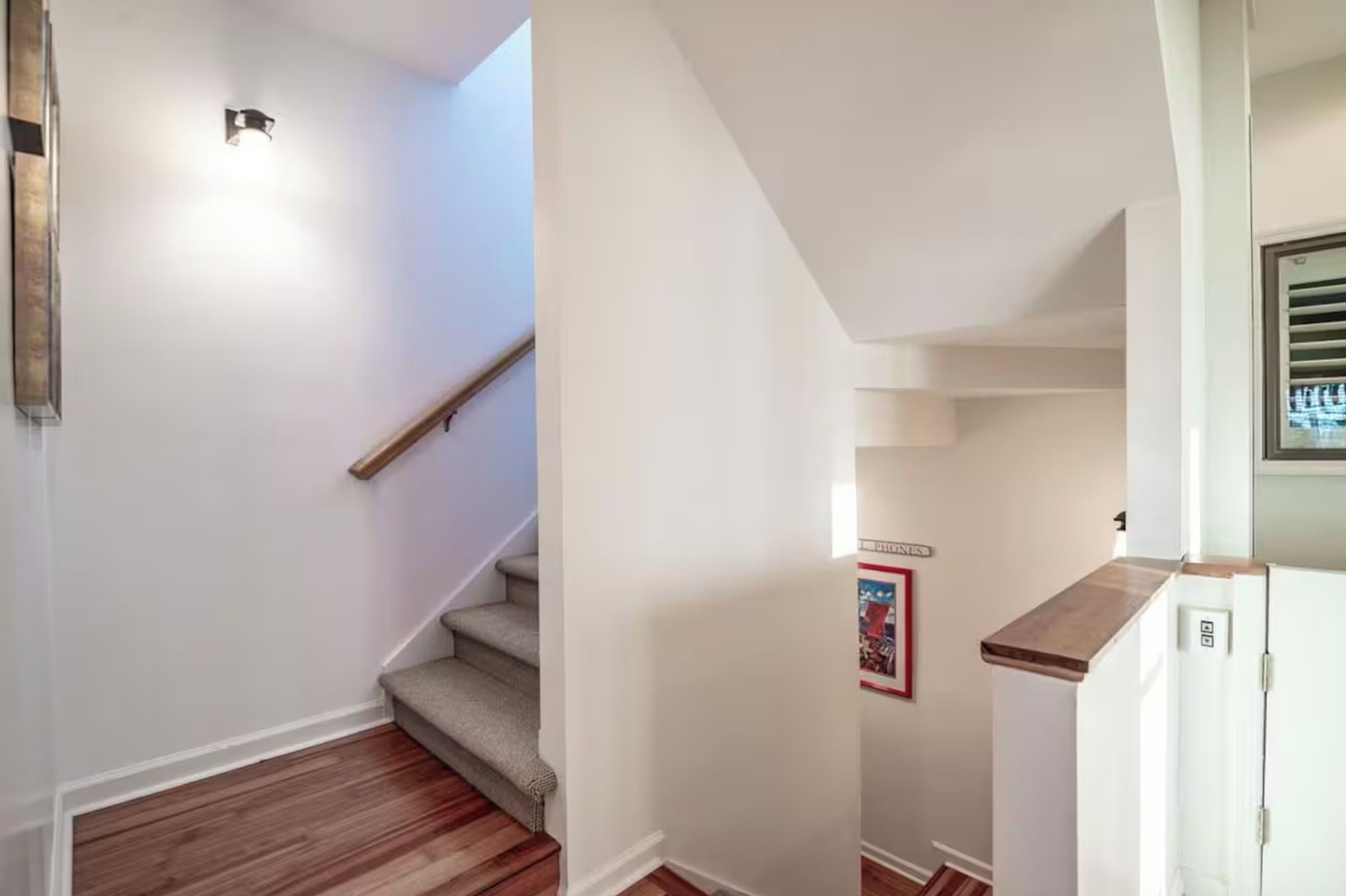 A staircase with a wooden handrail leading to a landing, a small kitchen area with a refrigerator, and a wall-mounted light fixture.