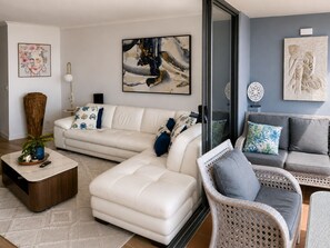 Ein modernes Wohnzimmer mit einem Ecksofa, einer Chaiselongue und einem Korbstuhl. Dekorative Kissen, eine Pflanzvase und Wandbilder sind vorhanden.