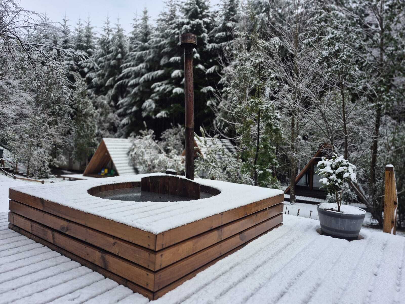 A wooden hot tub surrounded by snow-covered trees and a cabin.