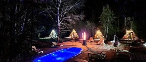 A nighttime outdoor area with a swimming pool, lounge chairs, and a fire pit.