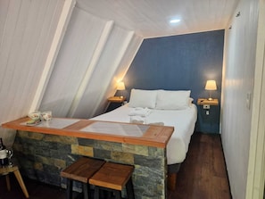 A room with a sloped ceiling, a bed with white bedding, two bedside lamps, a small wooden table with a chair, and a stone accent wall.