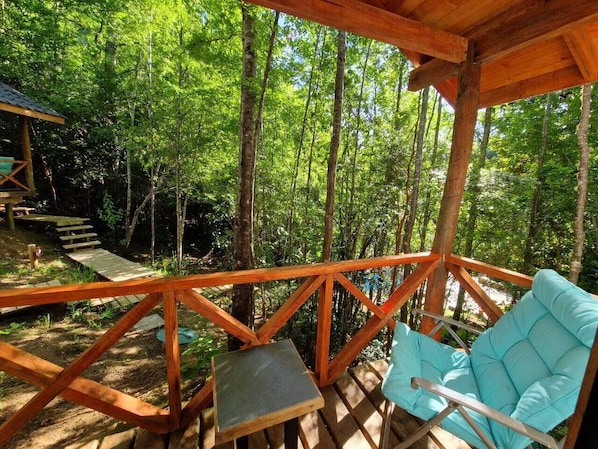 A wooden deck with a blue cushioned chair and a small table, surrounded by a forest. - Ecos del Manantial (Villarrica)