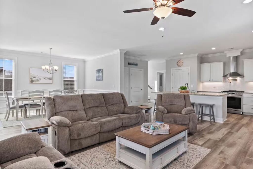 Ein modernes Wohnzimmer mit Sofa, Sessel und Couchtisch, ein Essbereich mit Stühlen und Tisch sowie eine Küche mit Schränken und Geräten.
