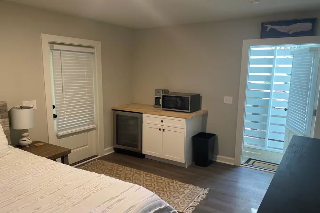 A bedroom with a bed, a bedside table, a small cabinet, a microwave, and a door with blinds.