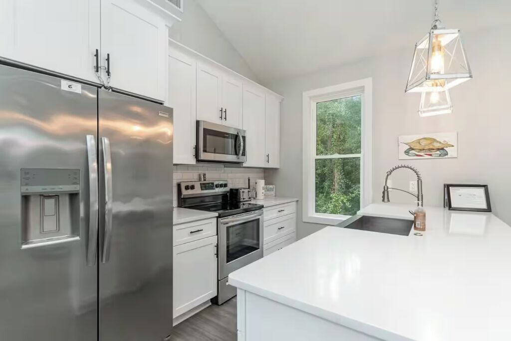 A modern kitchen with stainless steel appliances, white cabinetry, and a large window providing a view of greenery.