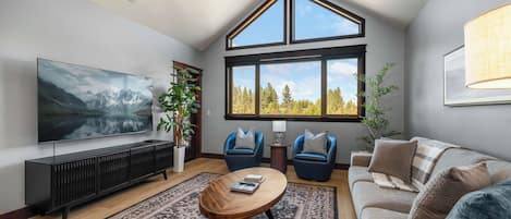 Sunlit loft living room with soaring vaulted ceiling, floor-to-peak picture windows, flat-screen TV, and plush sofa set — the view does all the talking.