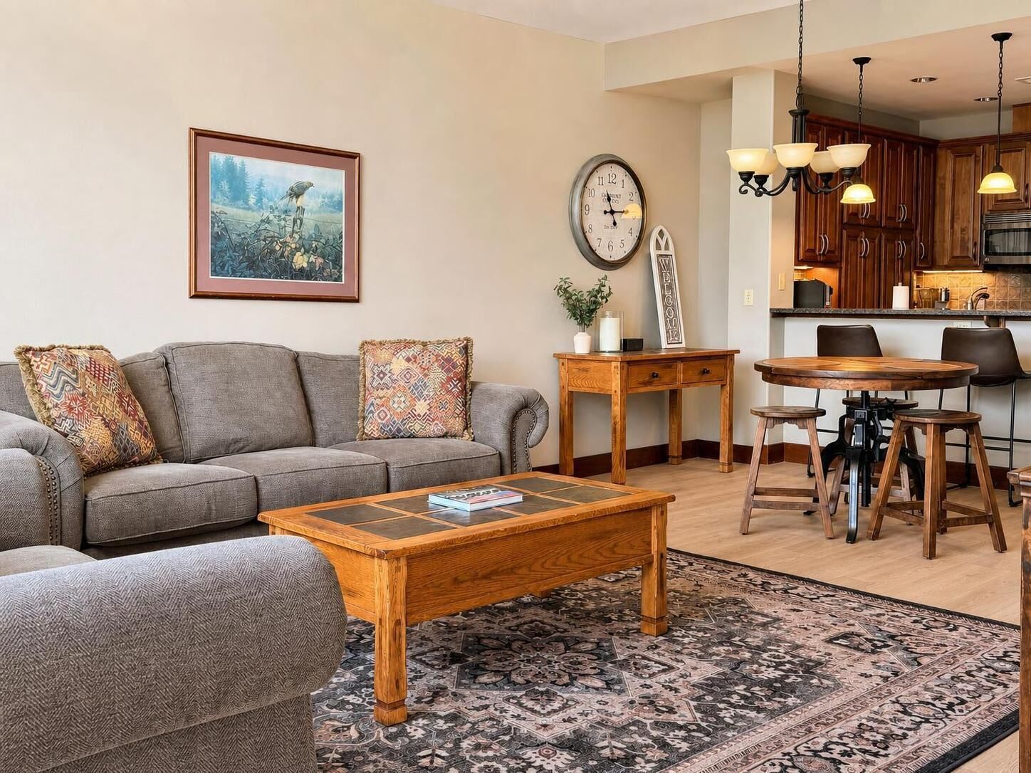 Ein Wohnzimmer mit Sofa, Couchtisch und Teppich. Es gibt einen Essbereich mit Tisch und Stühlen. An der Wand befinden sich eine Uhr und ein Willkommensschild.