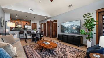 A modern living room with a large TV, a coffee table, and a sofa. There is a kitchen area in the background with wooden cabinets and a dining table.